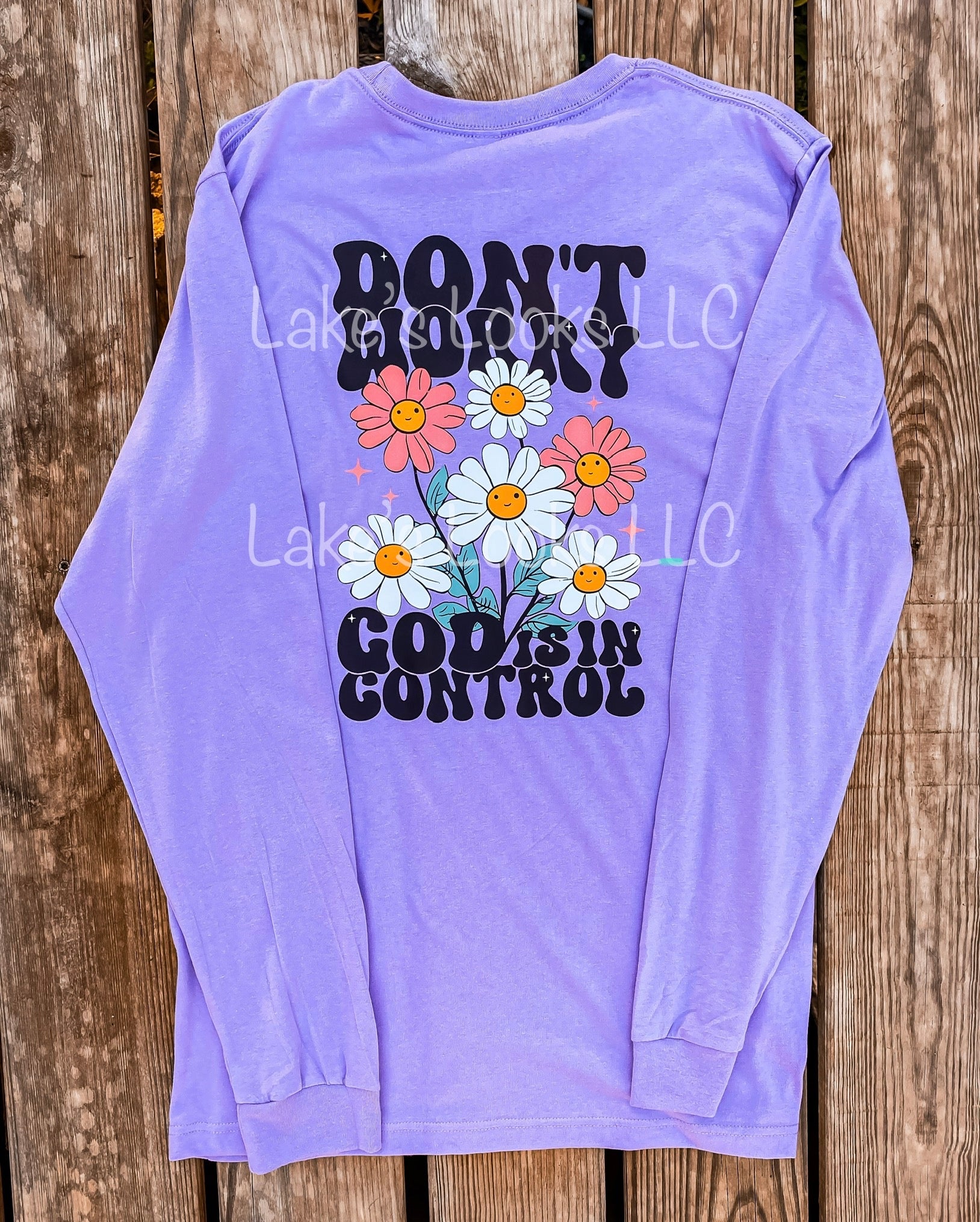 Retro religious graphic tee with pink and white flowers. God is in control. Featured on a purple long sleeve tee, this can be altered. 